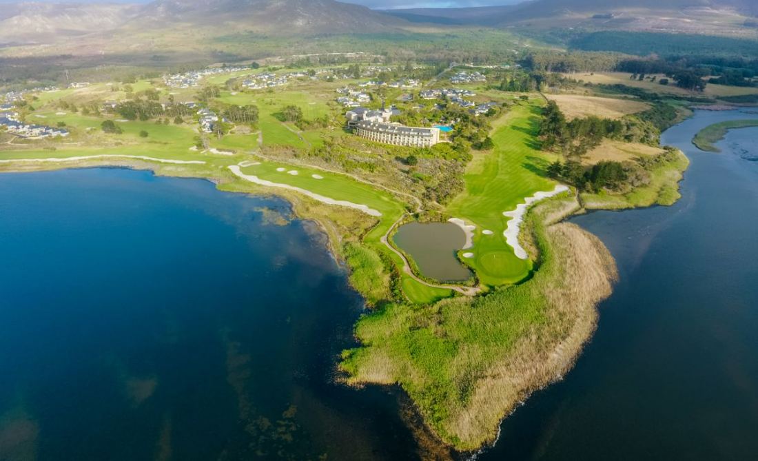 An aerial view of a golf course on a green peninsula surrounded by the deep blue water of a lagoon