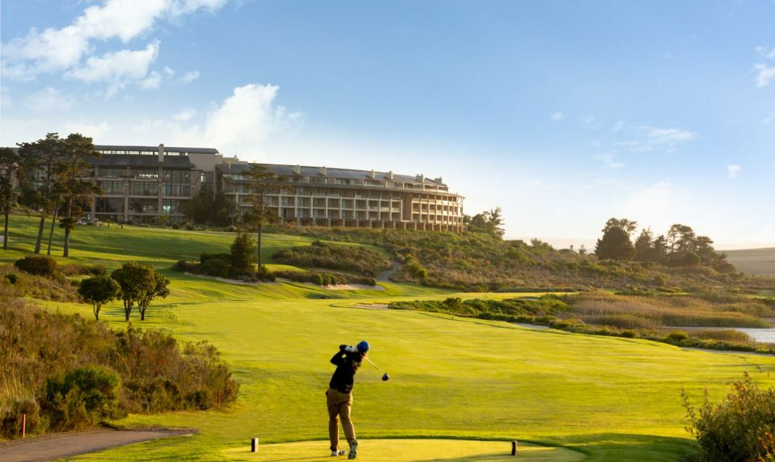 A golfer teeing off on a green fairway with the hotel on the rise beyond