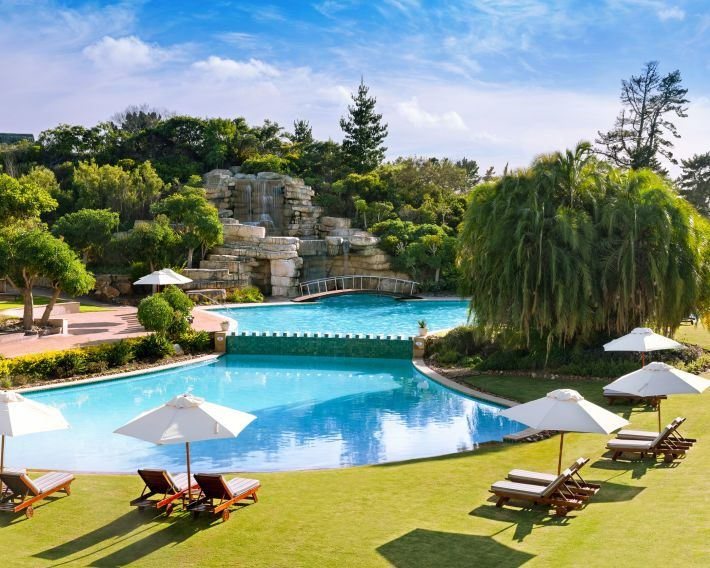 A swimming pool with umbrellas and loungers beside a rocky waterfall and willow trees