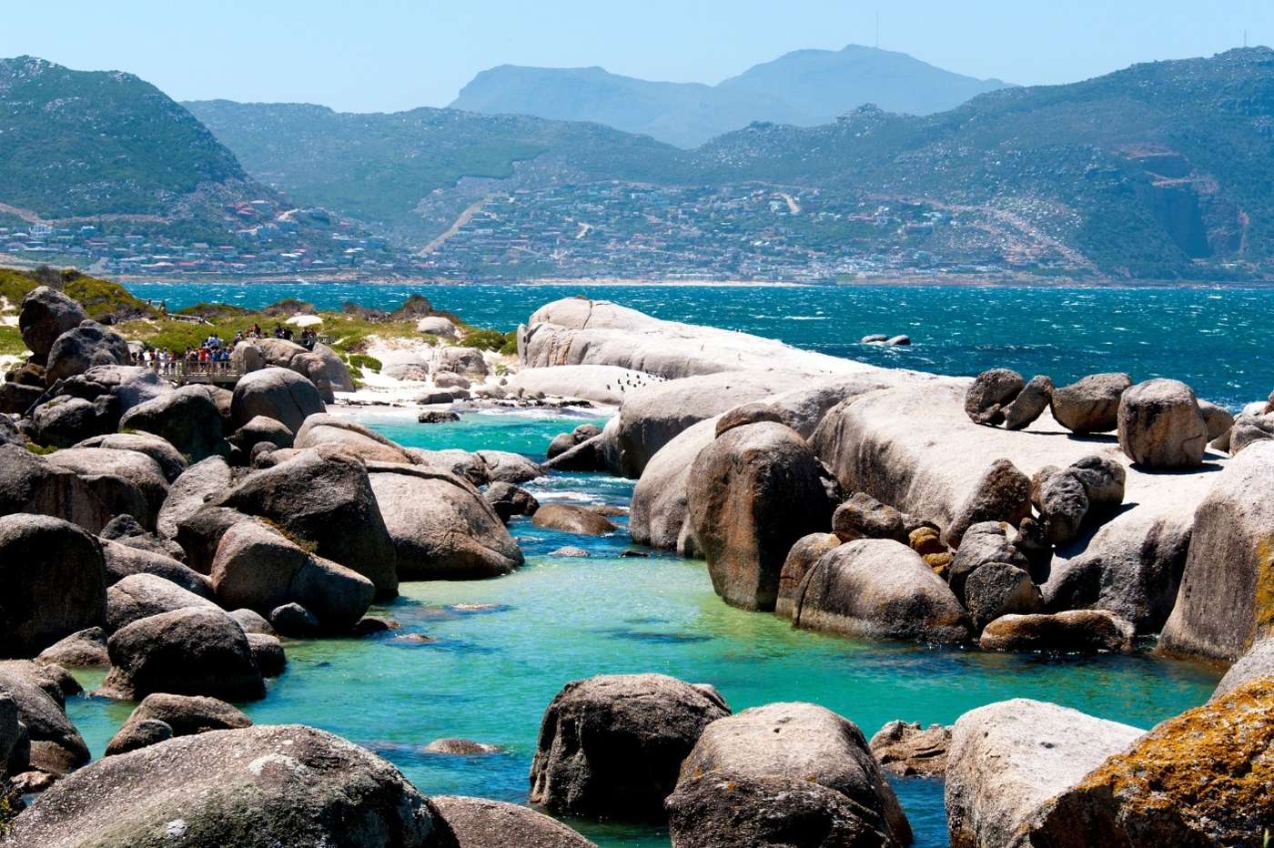 Turquoise water winding between great granite boulders, with the boardwalk, Simon's Town and the mountains beyond