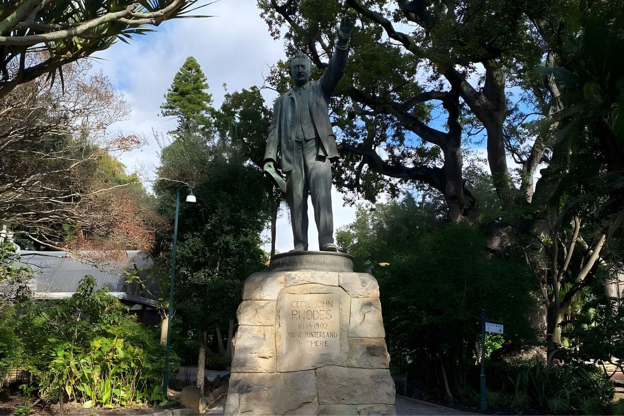 The bronze statue of Cecil John Rhodes on a stone plinth, his arm raised, among the trees of the garden