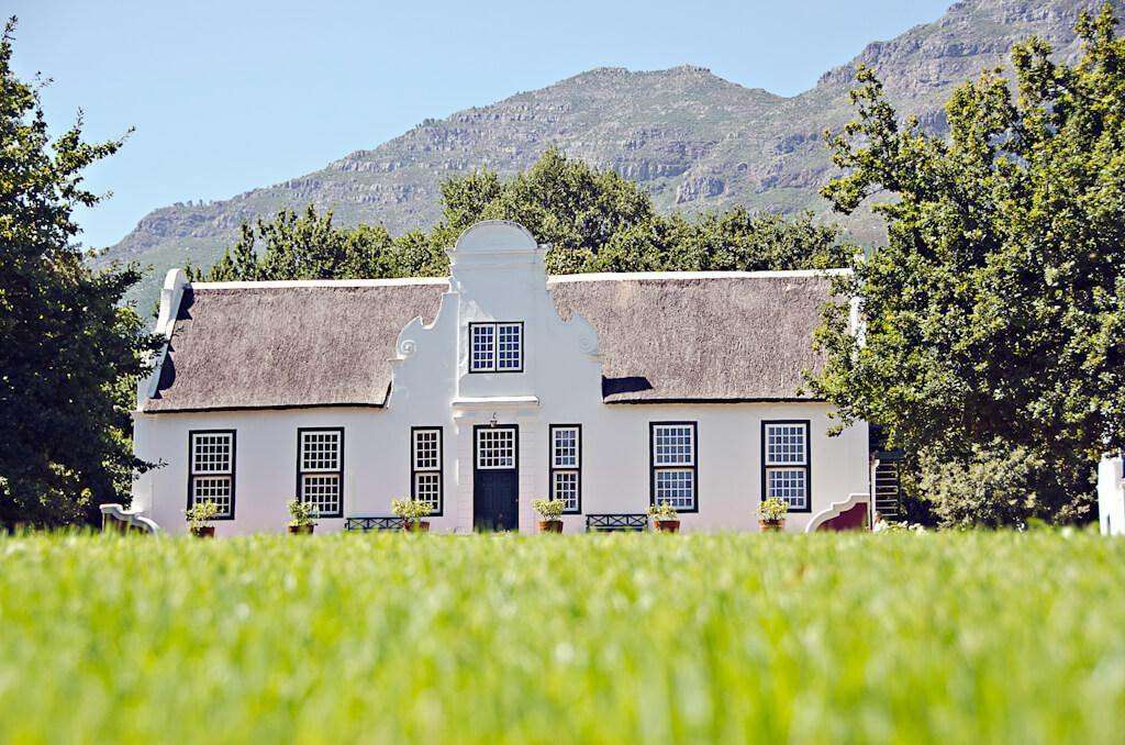 A white Cape Dutch manor house with a curved gable and thatch roof, mountains behind
