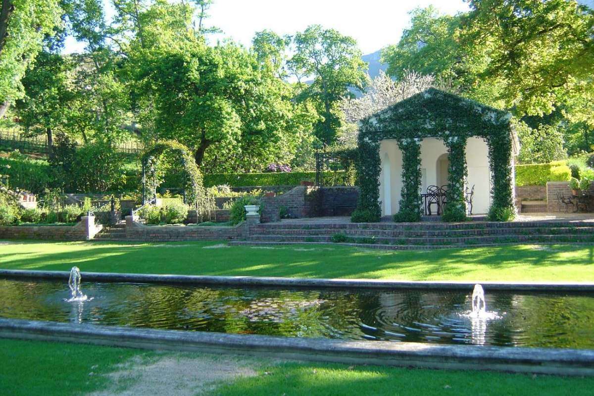 A long formal lily pond with fountains in front of a vine-covered pergola