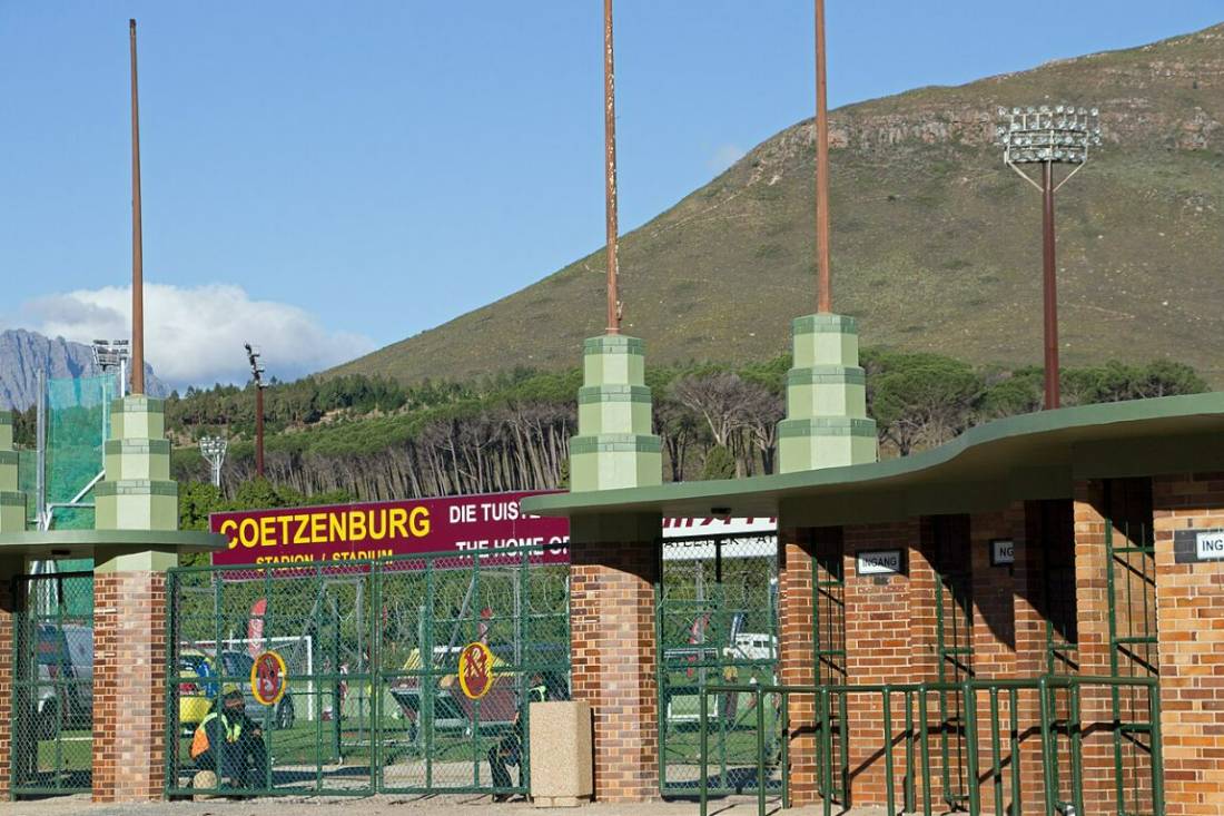 The entrance gates of a sports stadium below a green mountain