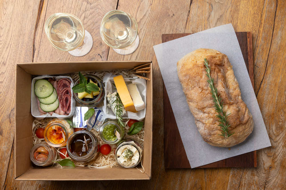 A picnic box with cheese, cold meats and preserves beside a loaf of bread and two glasses of white wine